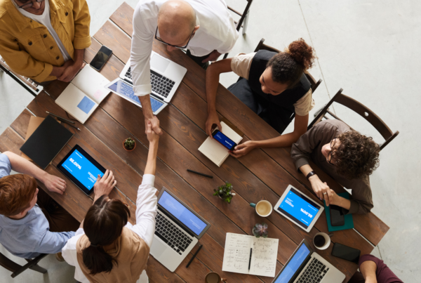 Overhead view of a diverse team gathered around a meeting table with laptops and tablets, two people shaking hands — illustrating the collaborative workplace practices that Equileap measures through its human capital data framework.